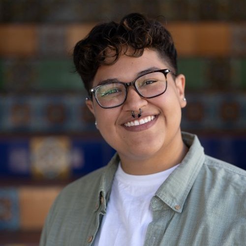 Person with short curly hair and glasses smiling, wearing a light green shirt over a white tee; colorful blurred background.