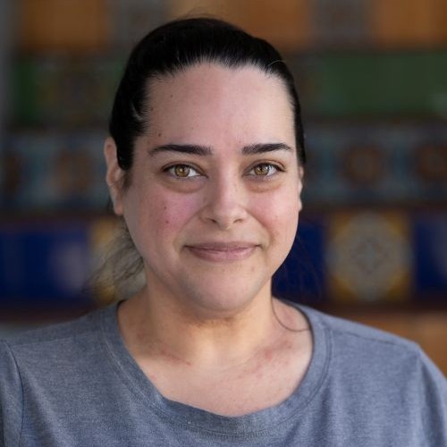 Close-up portrait of a smiling woman with dark hair tied back, wearing a gray shirt, blurred background.