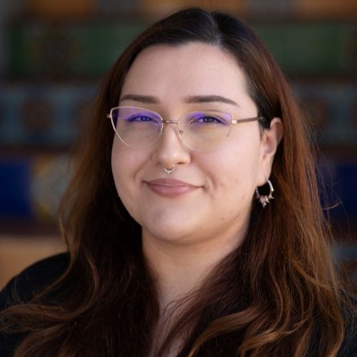 Portrait of a smiling woman with long brown hair, purple-tinted glasses, and a nose ring, against a blurred colorful background.