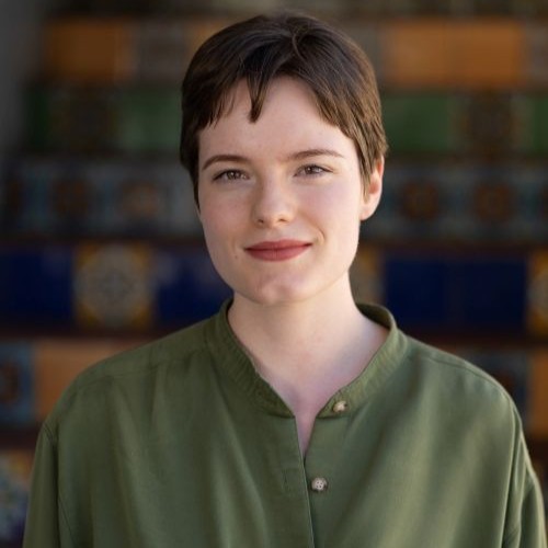 Head-and-shoulders portrait of a person with short brown hair, wearing an olive-green shirt, smiling softly against a blurred colorful background.