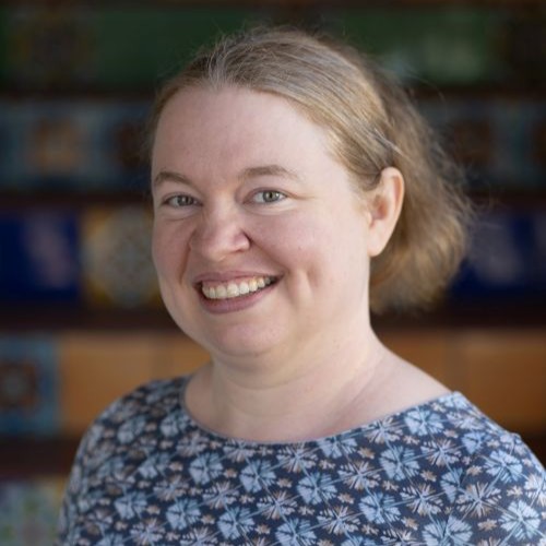 Smiling woman with light brown hair wearing a blue floral-patterned blouse, indoors with a blurred background.