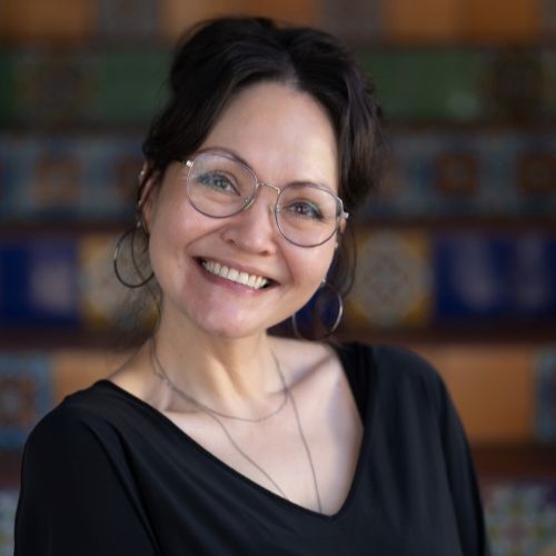 Portrait of a smiling woman with short dark hair and glasses, wearing a black top, in front of a colorful tiled background