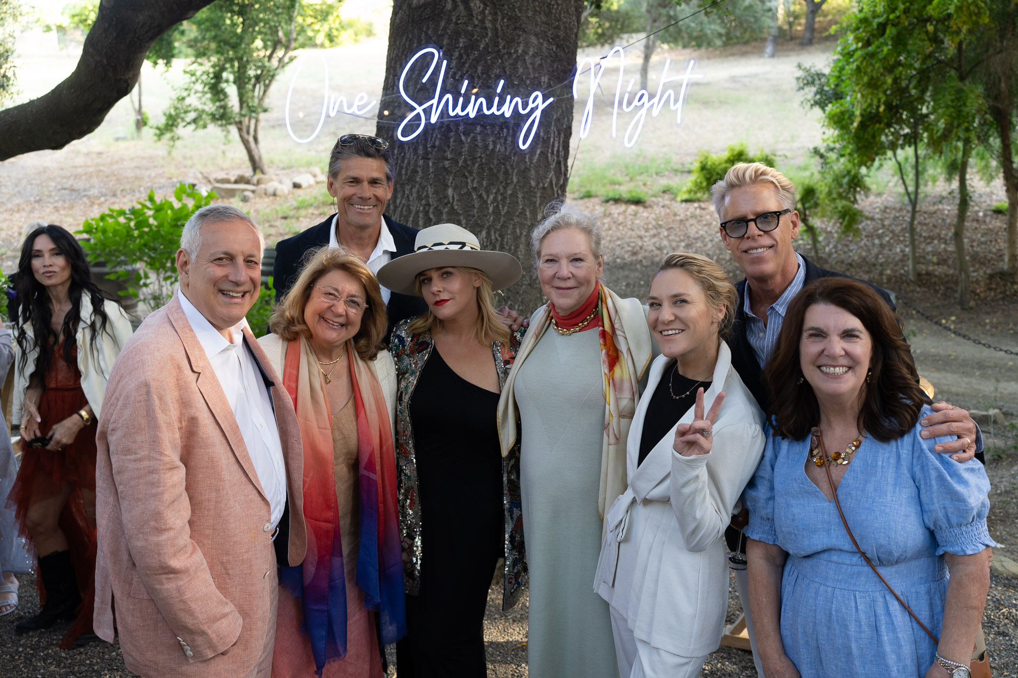 Group of smiling adults posing for a photo outdoors with a neon sign reading 'One Shining Night' in the background.