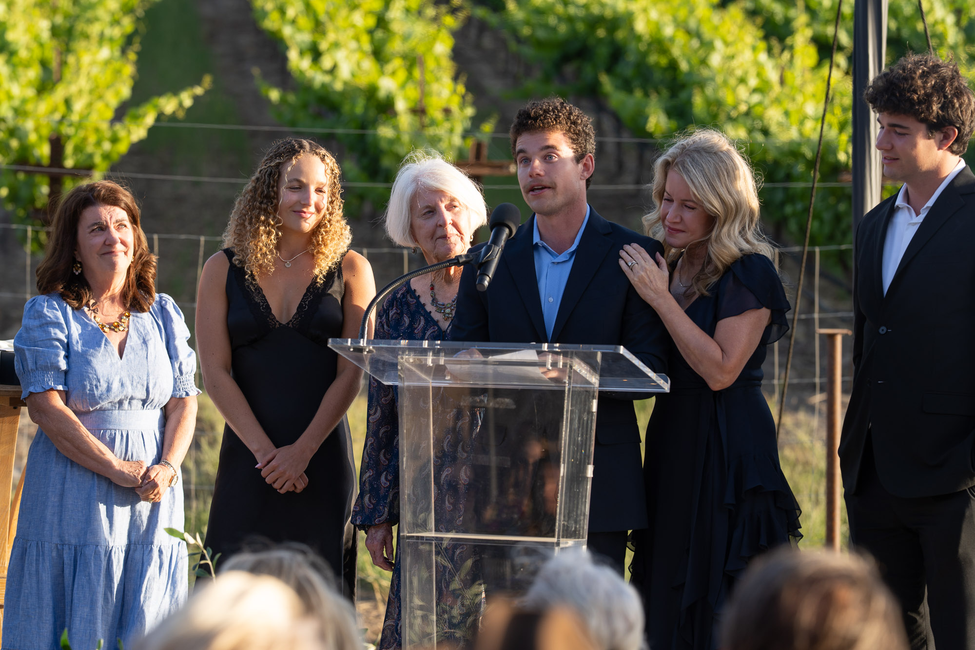 A group of six adults stands outdoors behind a clear podium as a man speaks into a microphone, with others listening attentively in formal attire against a sunny vineyard backdrop.