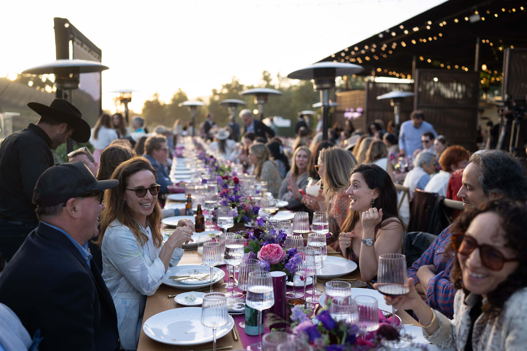 Outdoor evening banquet with guests seated along a long table, purple flower centerpieces and wine glasses under string lights.