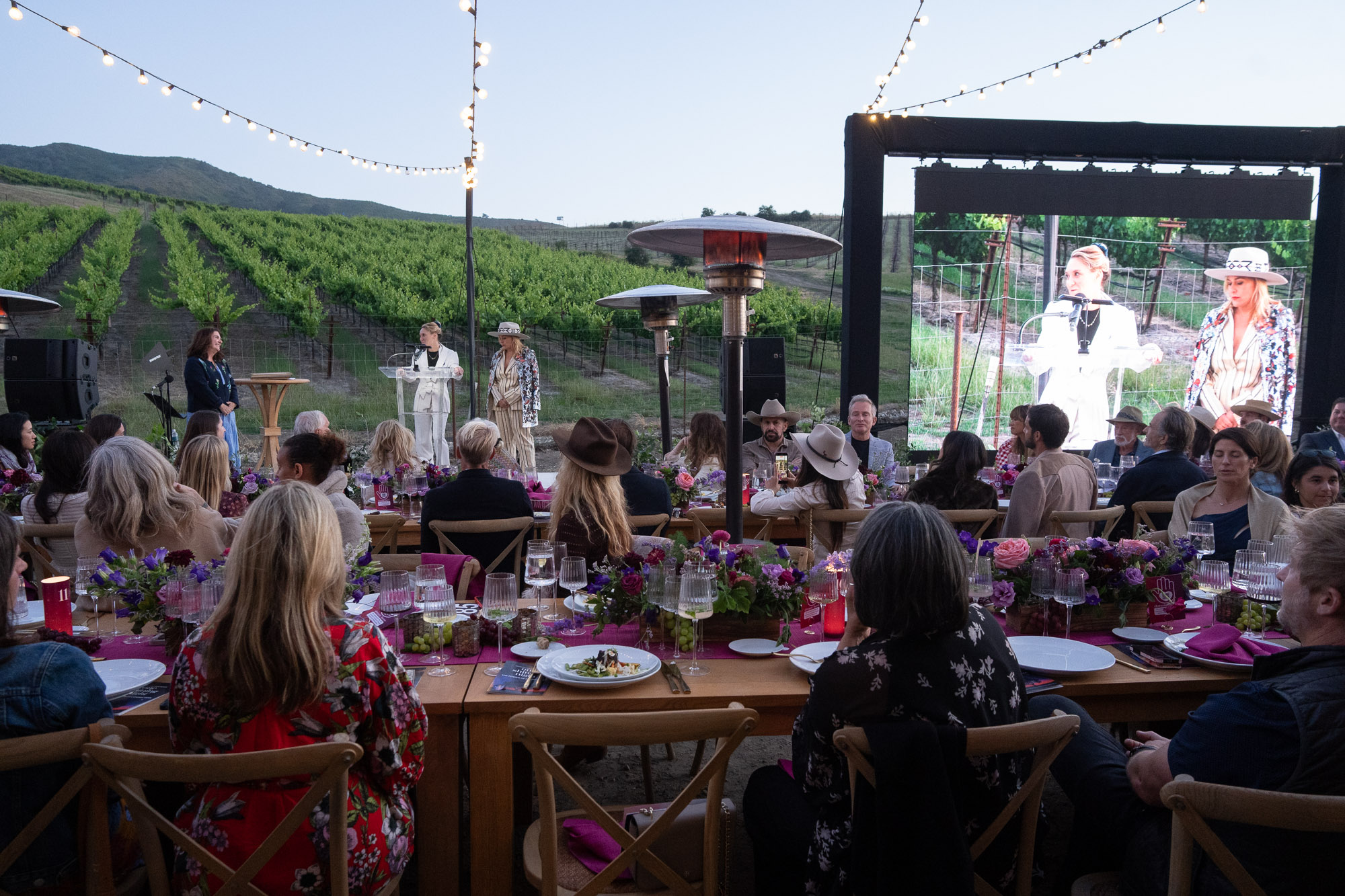 Outdoor vineyard event with guests seated at long tables, string lights overhead, and speakers on a stage with a large screen.