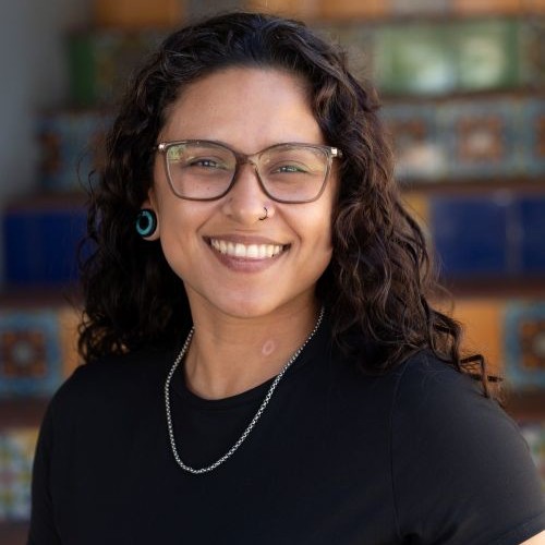 Smiling woman with curly hair and glasses, wearing a black top, nose ring, turquoise earrings, and a silver necklace; blurred colorful background.