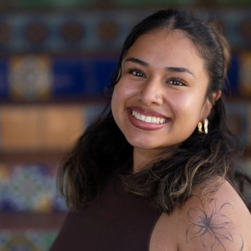 Smiling woman with dark hair, brown top, gold hoop earrings, nose stud, and a floral tattoo on her shoulder.