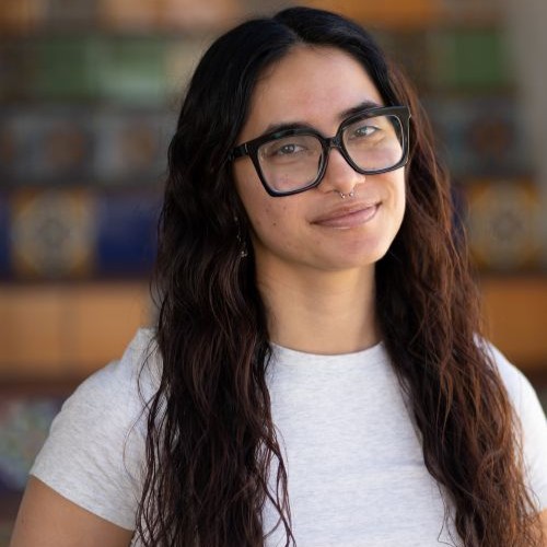 Portrait of a woman with long dark hair, black glasses, and a light gray T‑shirt, in a library setting.
