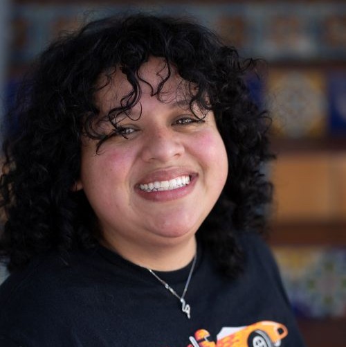 Smiling woman with curly black hair, wearing a black shirt and a necklace, in a warm indoor setting.
