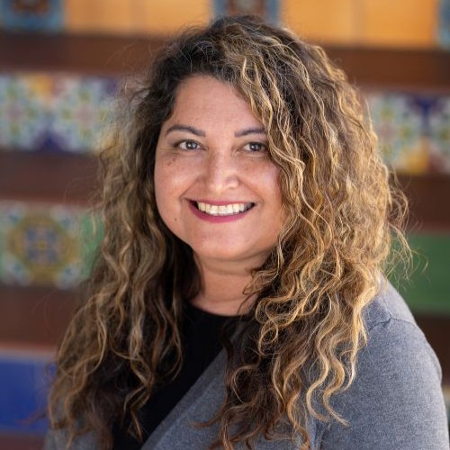 Portrait of a smiling woman with long curly hair wearing a gray cardigan, against a colorful blurred background.