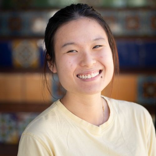 Smiling woman wearing a pale yellow shirt, posed for a portrait with a blurred colorful background.