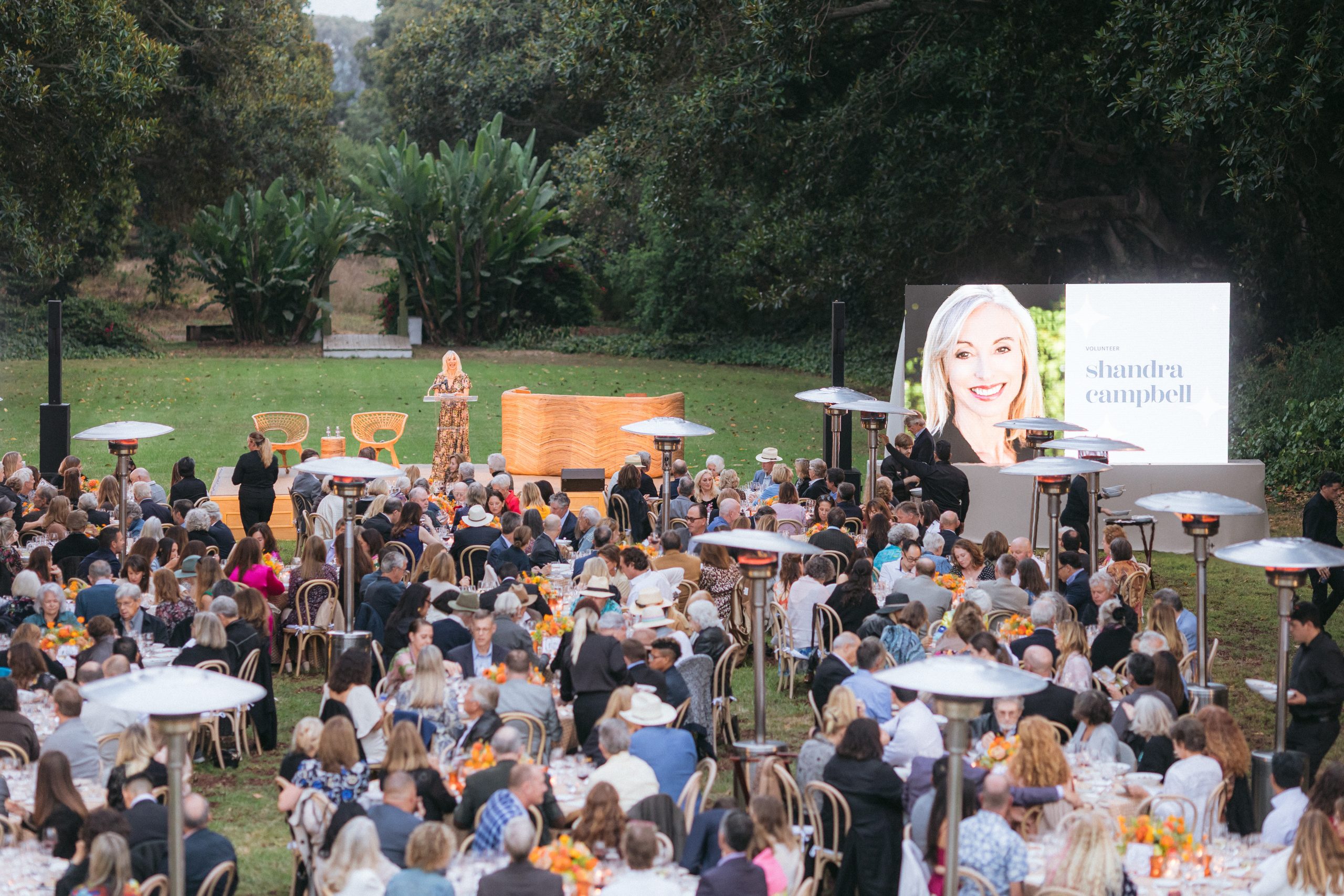 Outdoor gala in a park: large crowd seated at round tables facing a stage with an oversized screen showing a smiling woman in a bright photo, trees in the background.