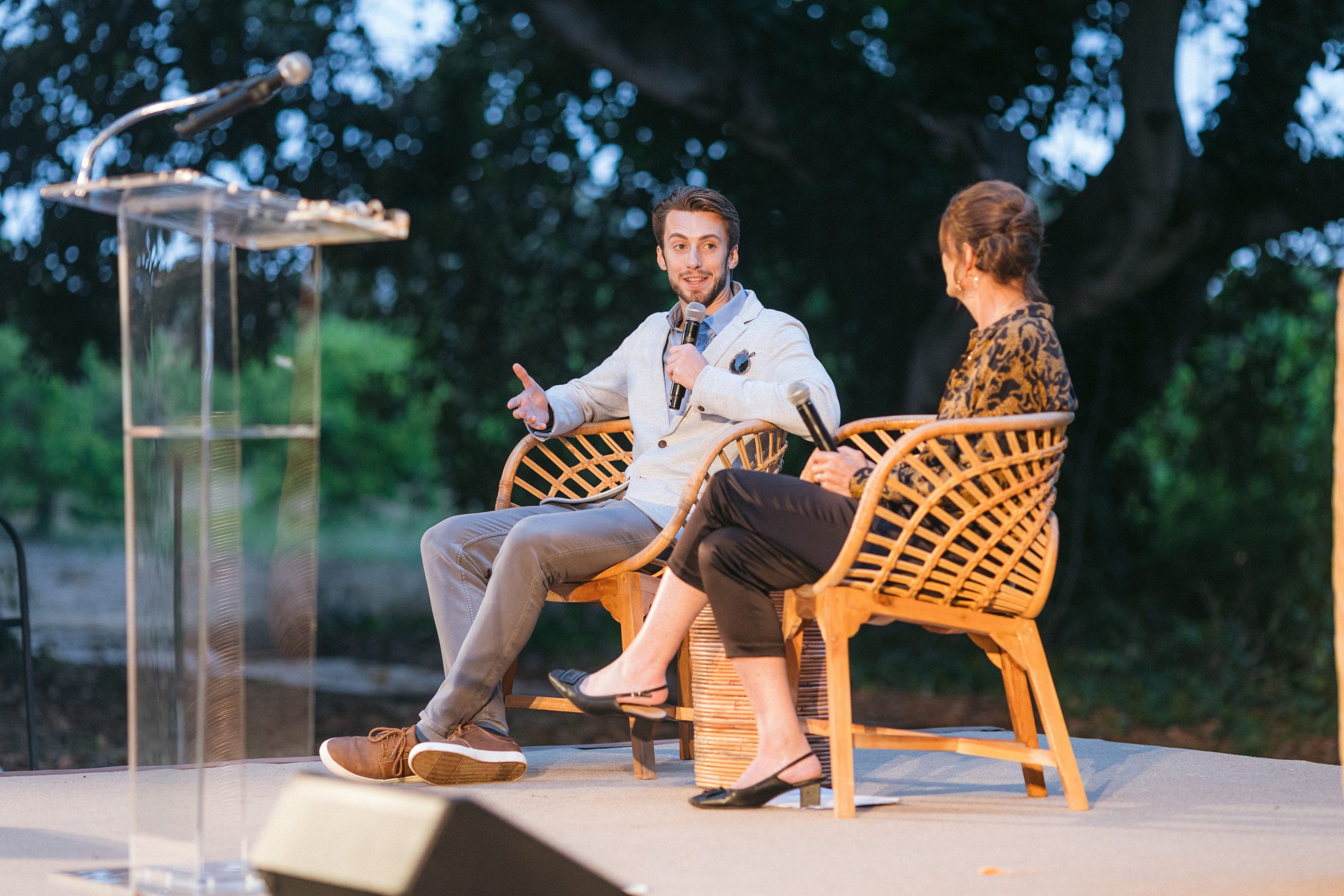 Two people sit in wicker chairs on an outdoor stage, speaker with microphone talking to a woman audience member beside him.