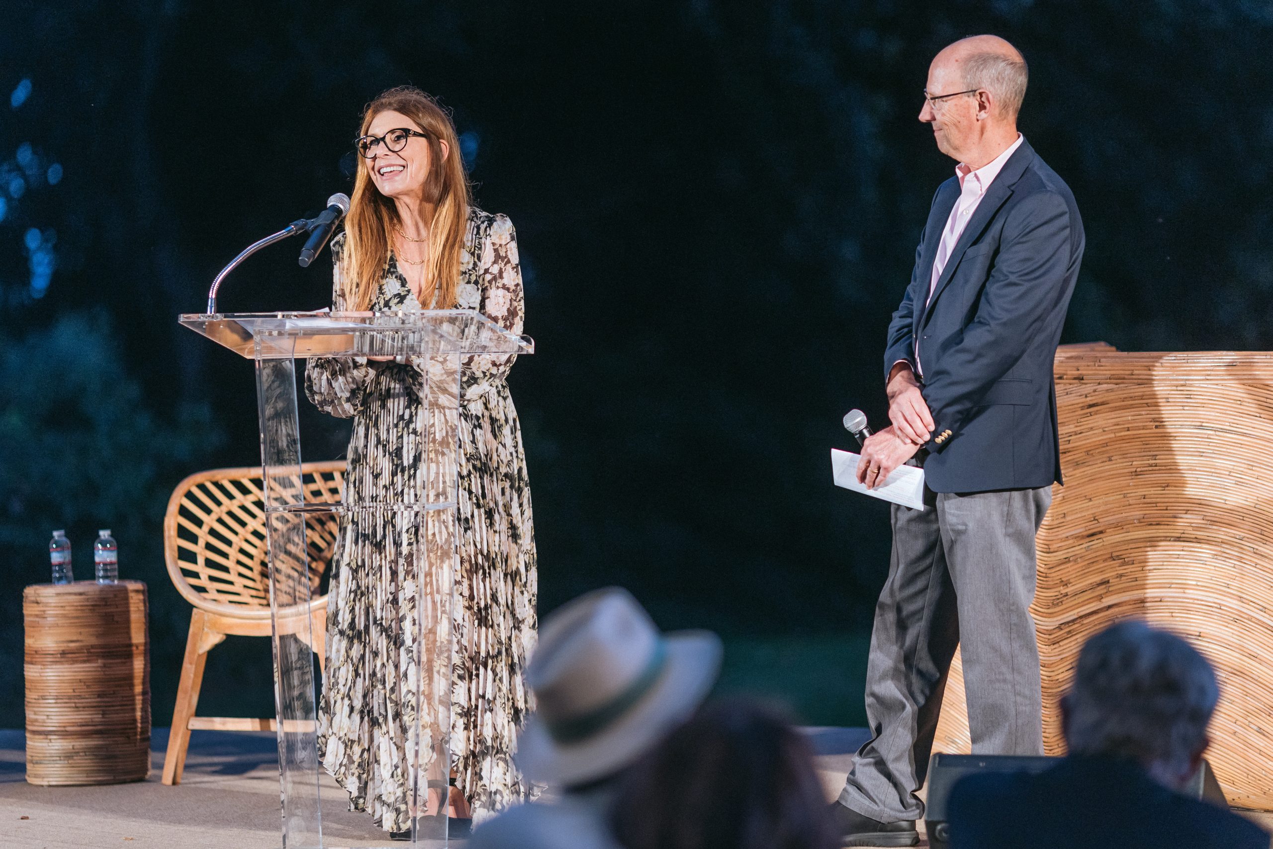 Woman in a patterned dress speaks at a clear lectern with microphone on stage, while a man in a suit stands nearby during an outdoor event.