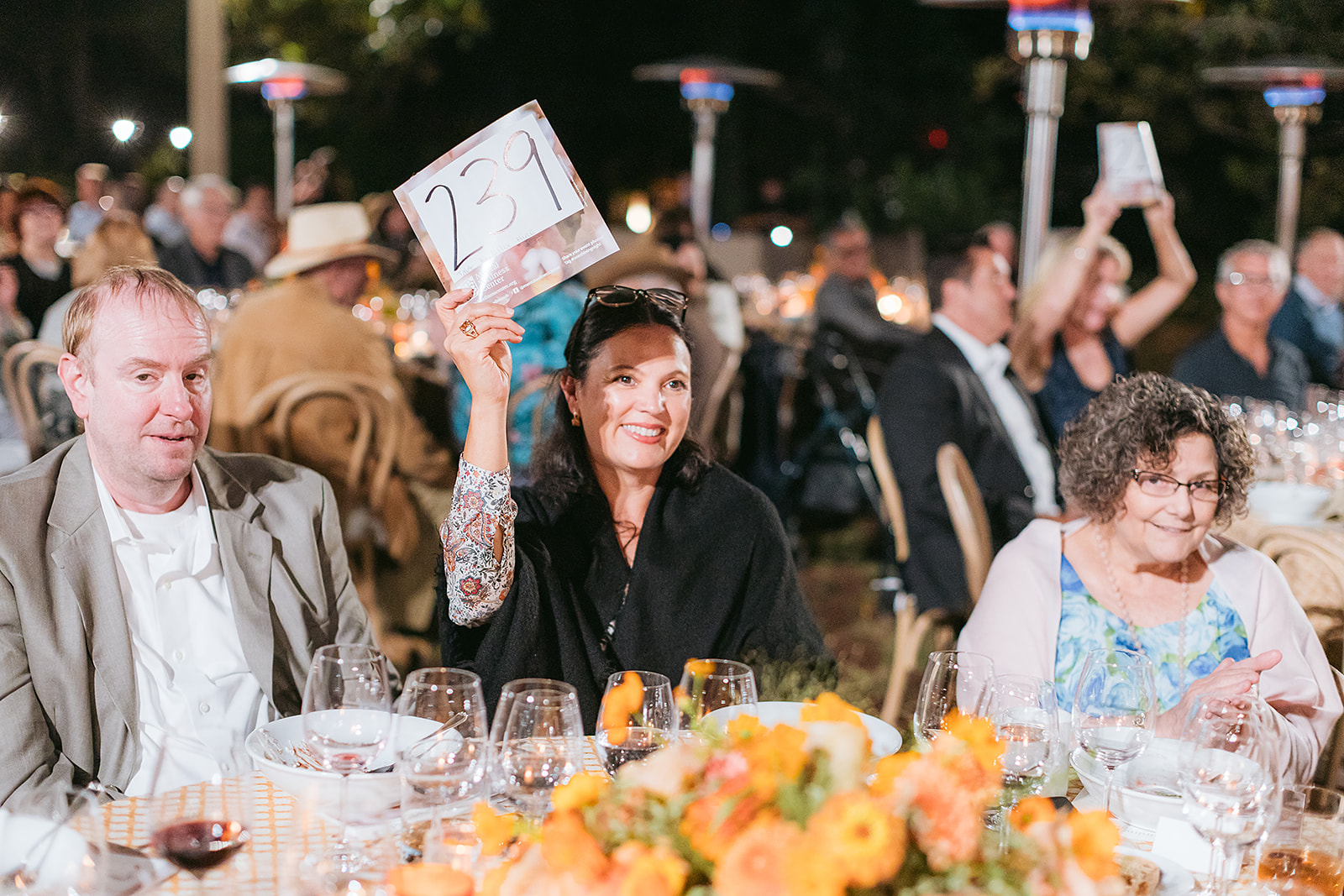Woman at a dinner event raises a numbered placard reading '231' while seated at a table with glasses and flowers.