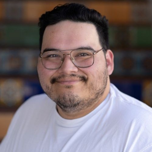 Smiling man with short dark hair and round glasses in a white shirt, blurred shelves behind him.