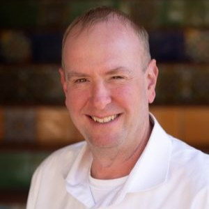 Smiling middle-aged man with short hair wearing a white polo shirt, outdoor background.