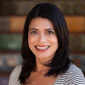 Portrait of a smiling woman with shoulder-length dark hair wearing a striped top, against a brick background.
